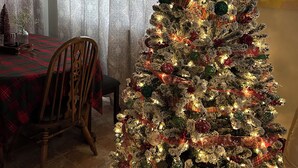 A Christmas tree decorated with lights and ornaments in a dining area with patterned curtains and a chandelier.