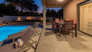 A poolside area with lounge chairs and a dining table, surrounded by a landscaped garden and a view of trees.