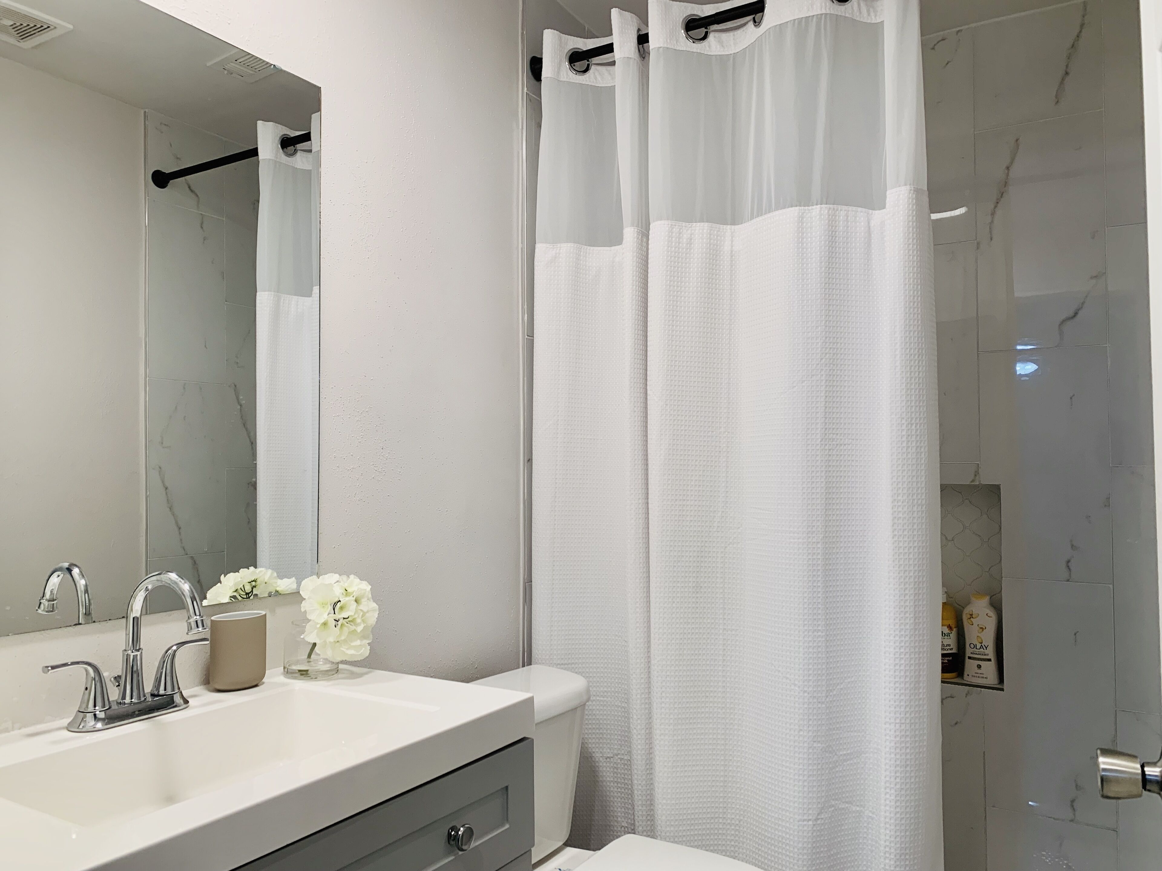 A bathroom with a white sink, mirror, and toilet. There is a shower curtain and a small shelf with toiletries.