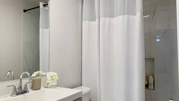 A bathroom with a white sink, mirror, and toilet. There is a shower curtain and a small shelf with toiletries.