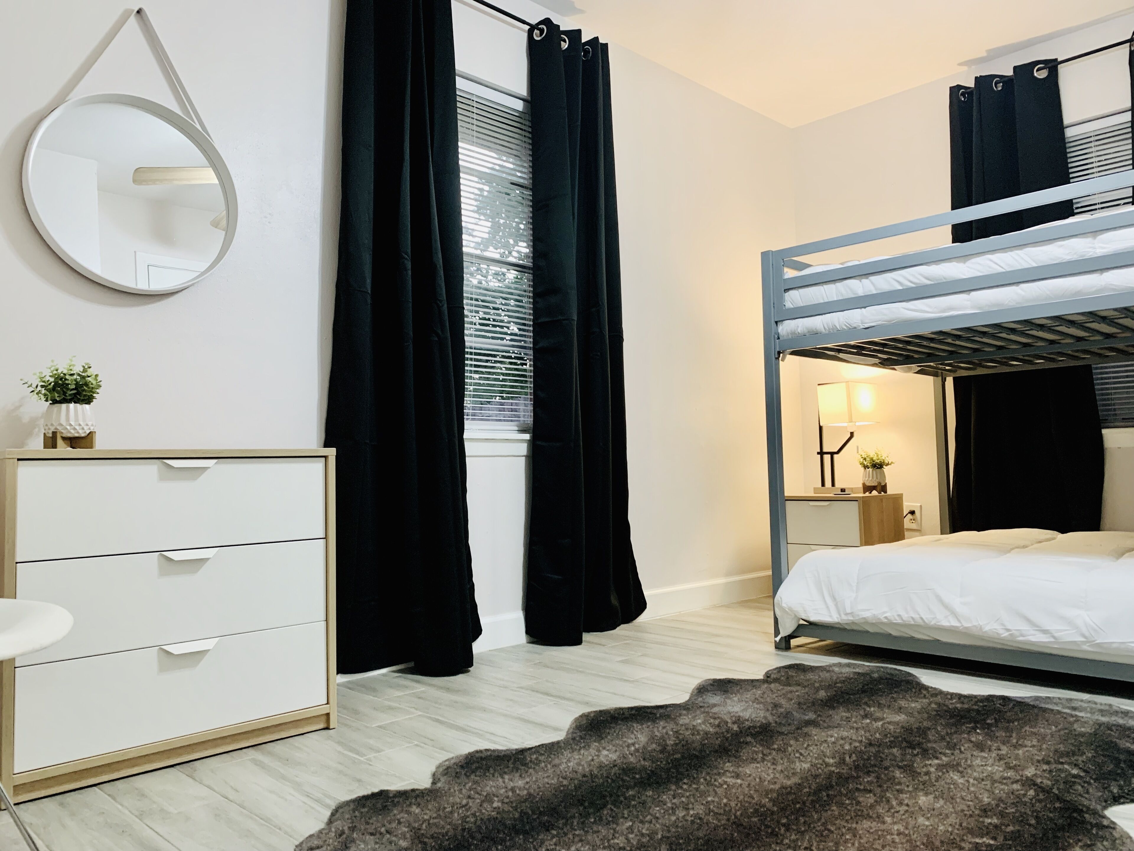 A bedroom with a bunk bed, a bedside table with a lamp, a dresser, and a round mirror on the wall.