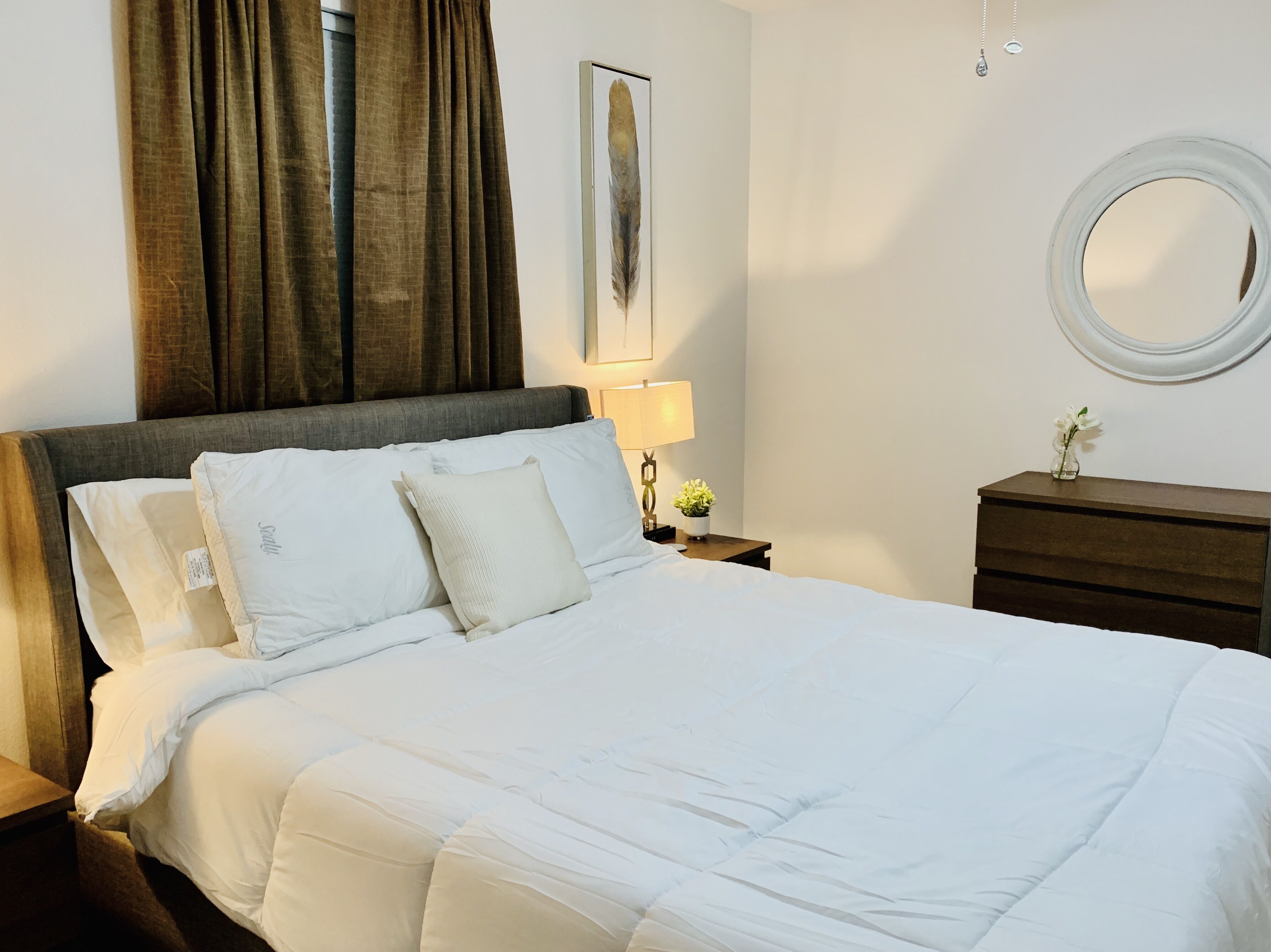 Una cama bien tendida con ropa blanca, una mesita de noche con una lámpara y una planta pequeña, un tocador con espejo y una ventana con cortinas.