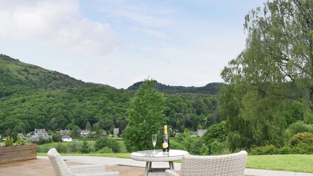 A wicker outdoor seating area with a table and chairs overlooking a lush green landscape and distant houses. - 1 bedroom accommodation in Aberfoyle (Aberfoyle)