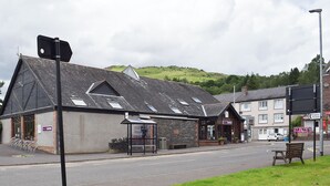 A building with a sloped roof, a public toilet sign, and a bench on the sidewalk. - 1 bedroom accommodation in Aberfoyle (Aberfoyle)