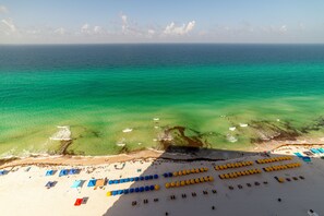 Une vue aérienne d’une plage avec des rangées de transats et de parasols, un océan bleu limpide et une plage de sable.