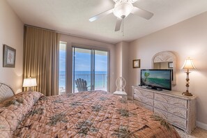 A bedroom with a bed, a television, a ceiling fan, a nightstand, a lamp, a chair, and a view of the ocean through a sliding door.