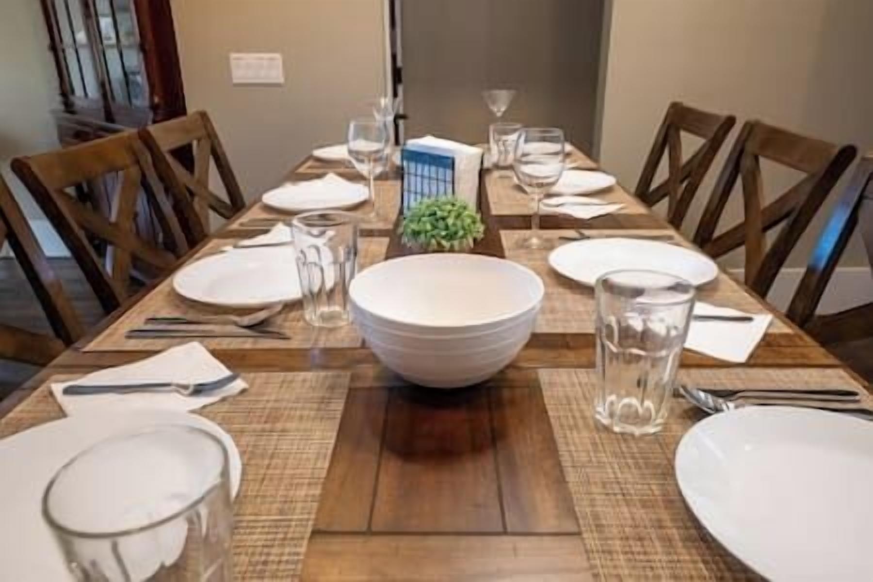 A dining table set for six with white plates, bowls, and glasses, surrounded by wooden chairs.
