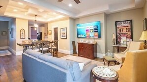 Een woonkamer met een bank, een salontafel en een televisie. Achtergrond toont een eethoek met een tafel en stoelen.
