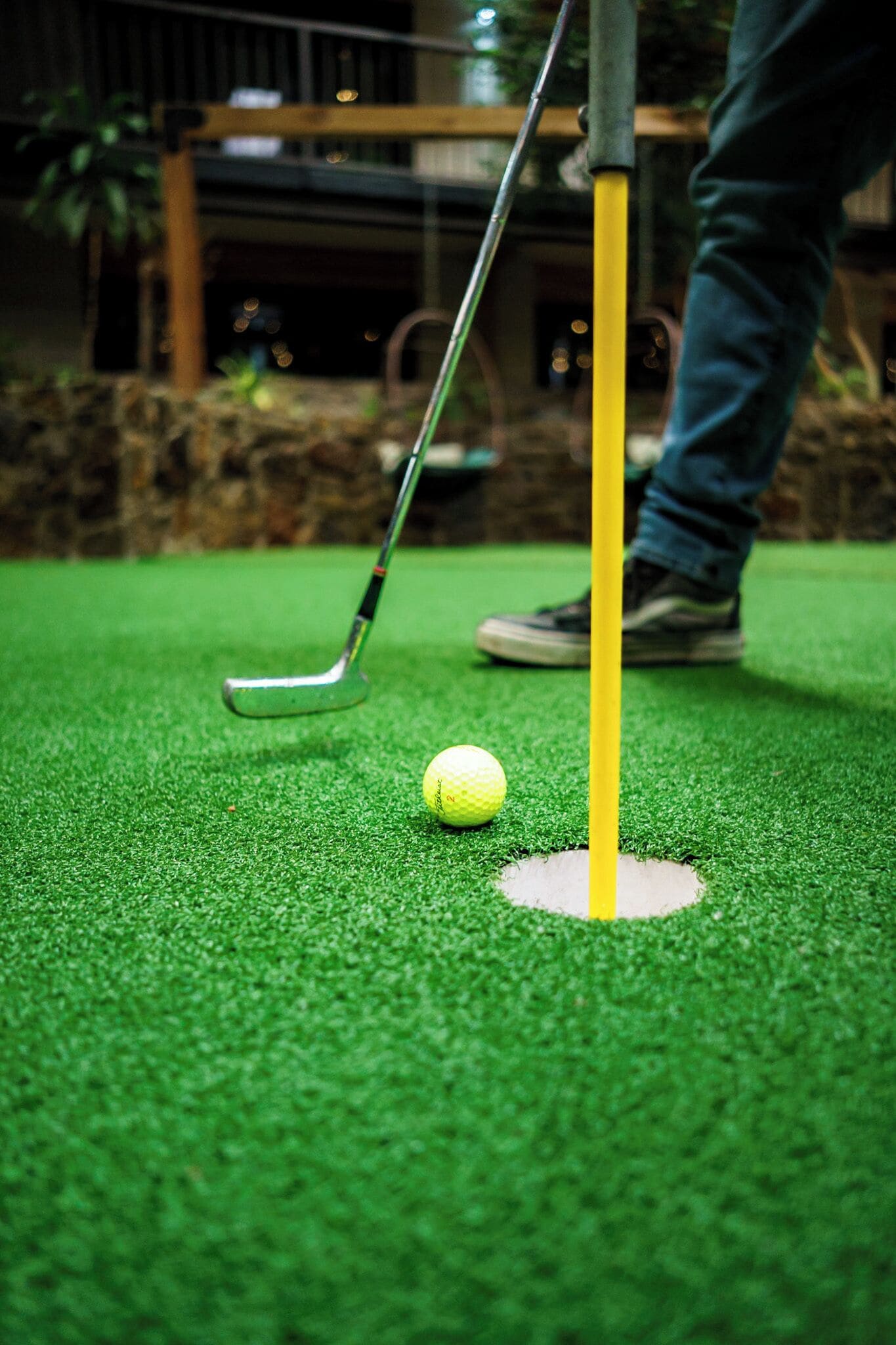 Una pelota de golf sobre césped artificial cerca de un hoyo con una bandera, y un pie de persona al fondo.