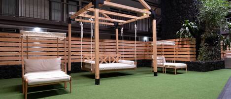 A wooden swing bed structure with a cushioned bench and a lounge chair in a room with artificial grass flooring and a stone wall backdrop.