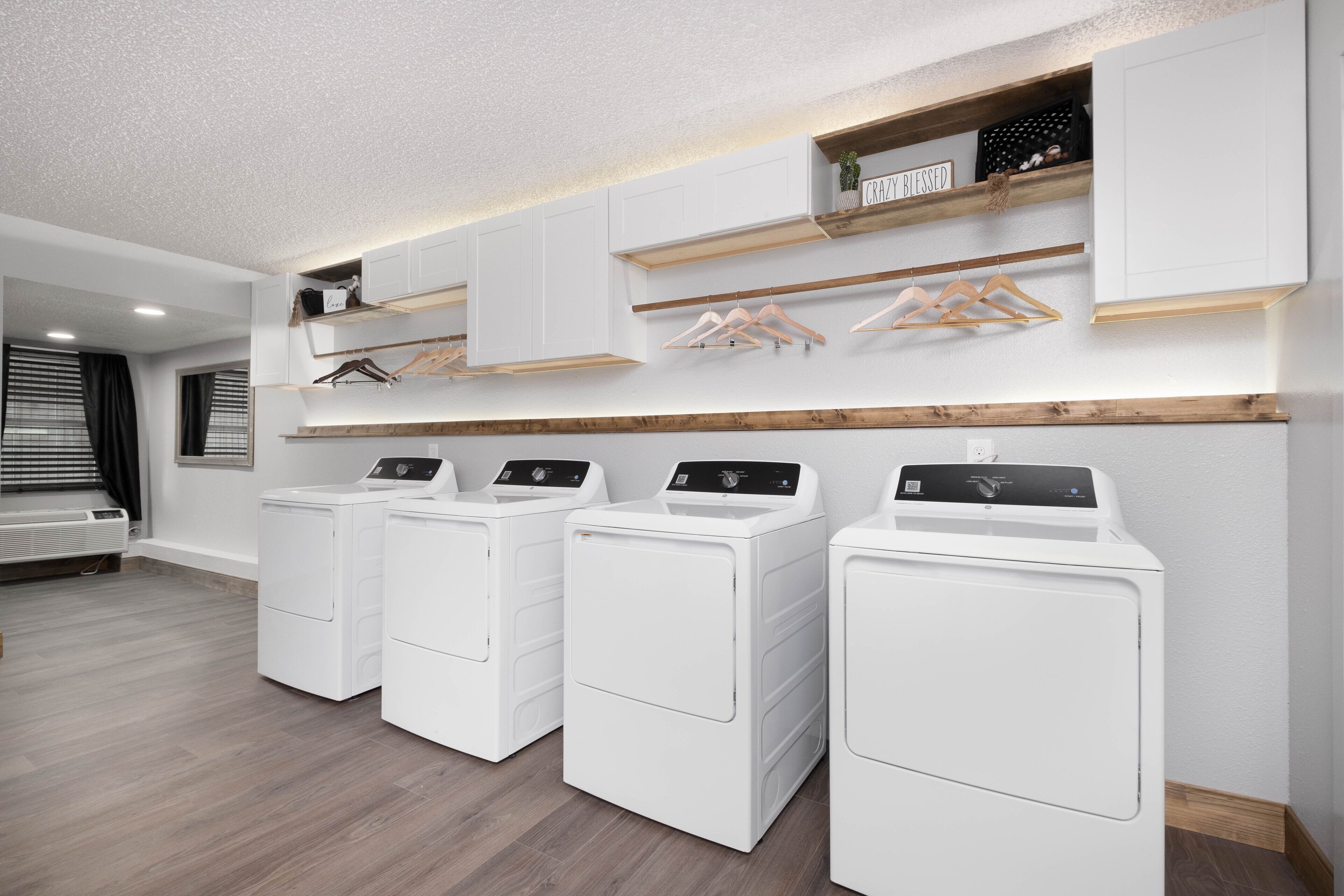 A laundry room with four white front-loading washers and dryers, wooden shelves above, and a window with blinds.