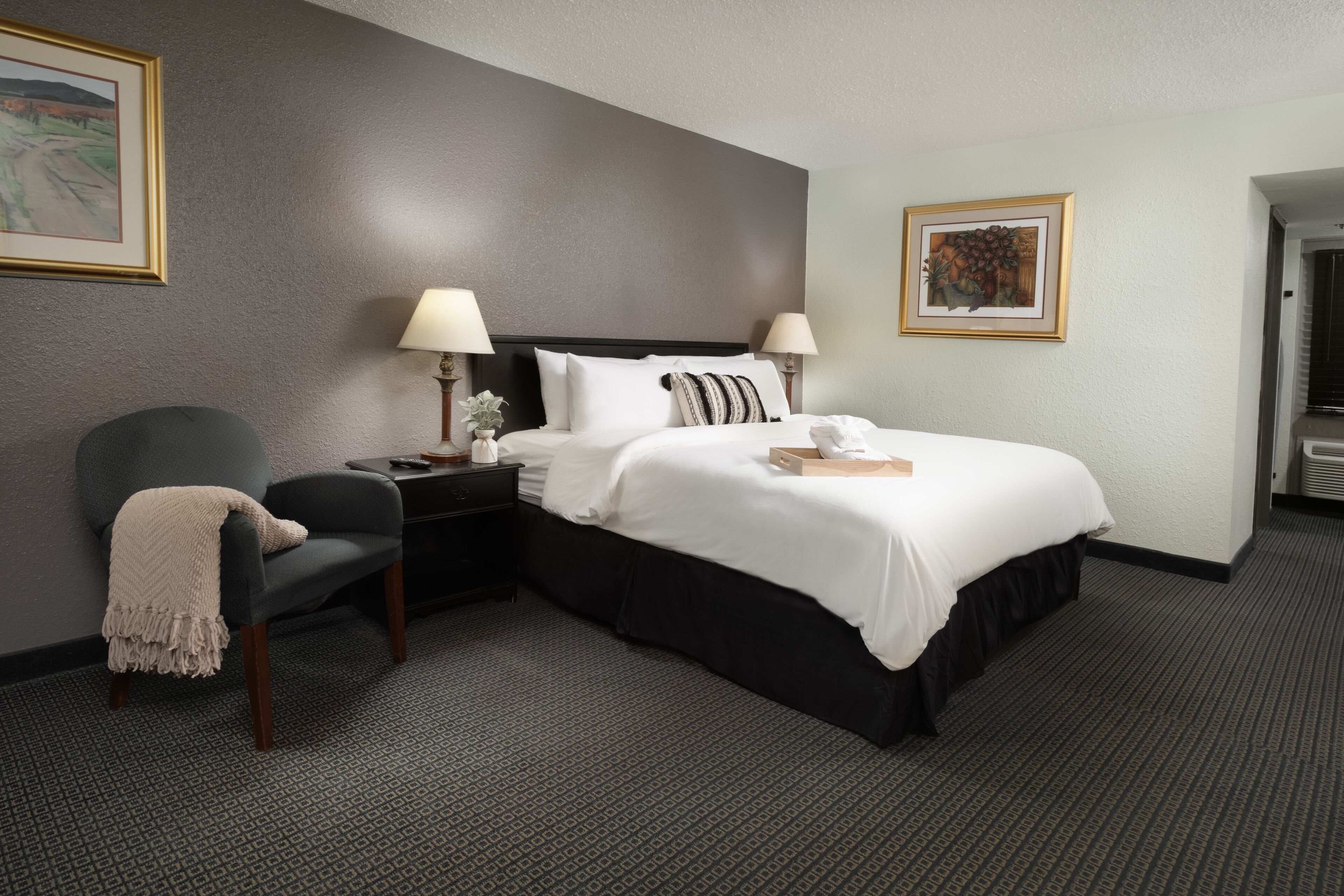 A hotel room with a bed, a chair, a nightstand, two lamps, and framed artwork on the walls.