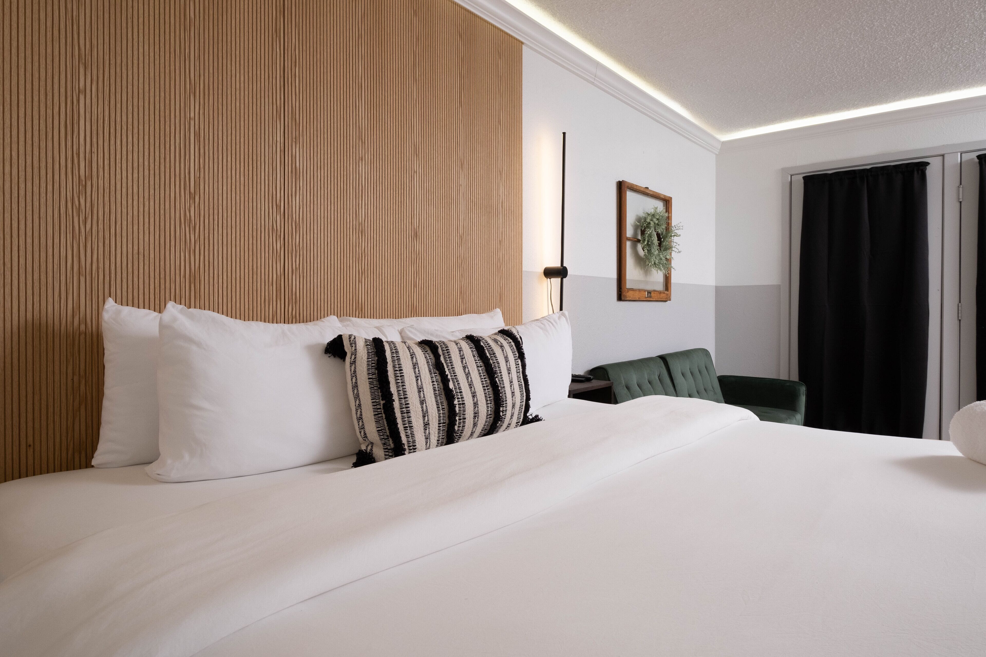 Ein Hotelzimmer mit einem großen Bett, einem Holzkopfende, einem grünen Sessel, einem schwarzen Vorhang und einem gerahmten Bild an der Wand.