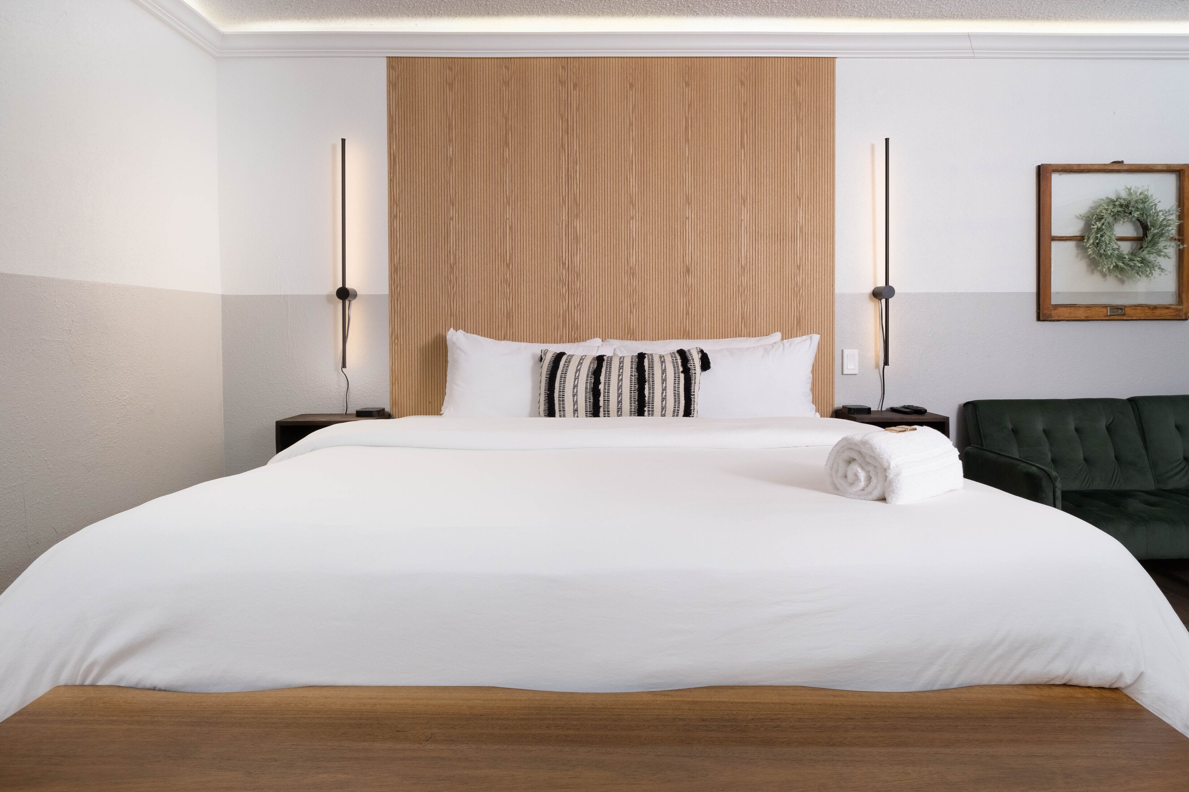 A hotel room with a large bed, wooden headboard, two bedside tables, a green sofa, and a wall-mounted wreath.