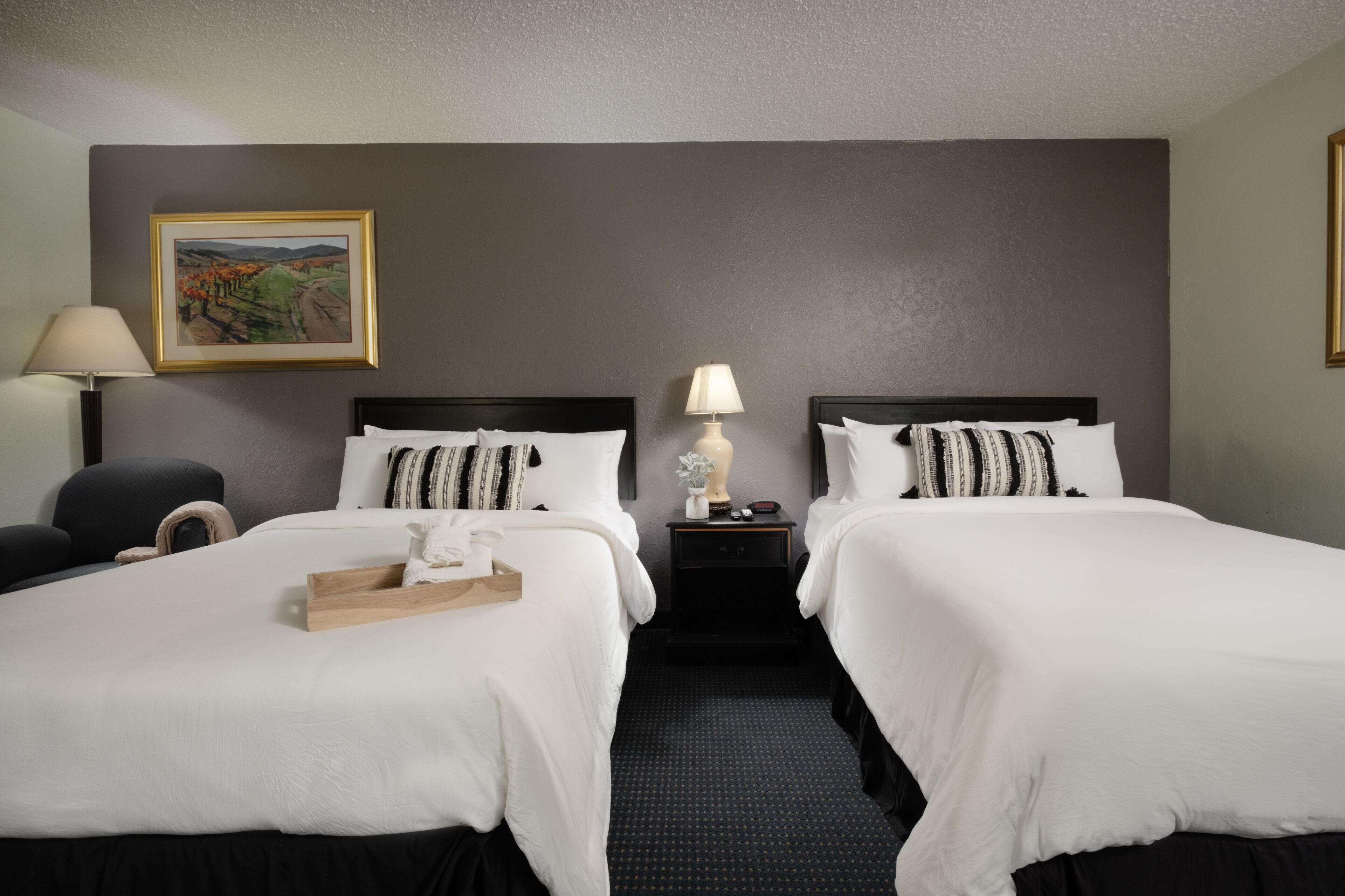 Habitación de hotel con dos camas, una mesita de noche, una lámpara, una silla y un cuadro en la pared.