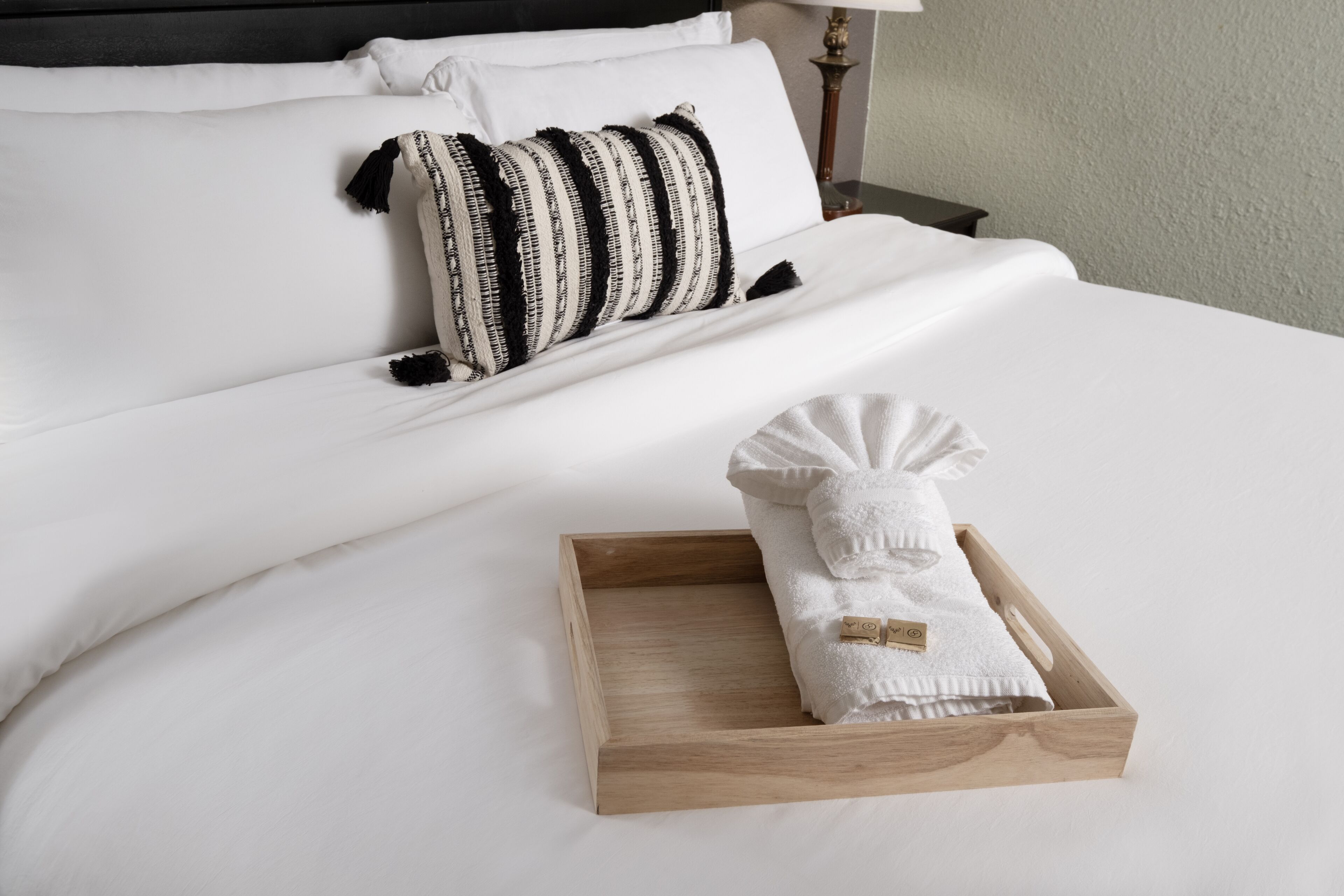A neatly made bed with white linens, a striped pillow, and a wooden tray with a folded towel.