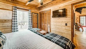 Un dormitorio tipo cabaña de madera con cama, ventilador de techo, televisor y ventana con cortinas.