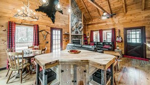 Interior rústico de una cabaña con una mesa de comedor de madera, sillas, una chimenea y una pared de piedra con monturas de animales.