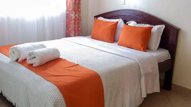 Una cama bien hecha con ropa de cama blanca y naranja, un cabecero de madera y una ventana con cortinas traslúcidas.