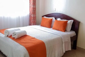 Een netjes opgemaakt bed met wit-oranje beddengoed, een houten hoofdbord en een raam met dunne gordijnen.