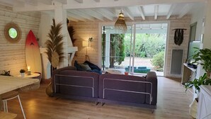 A modern living room with a wooden floor, a sofa, a dining area with a table and chairs, and a large window offering a view of the outdoors.