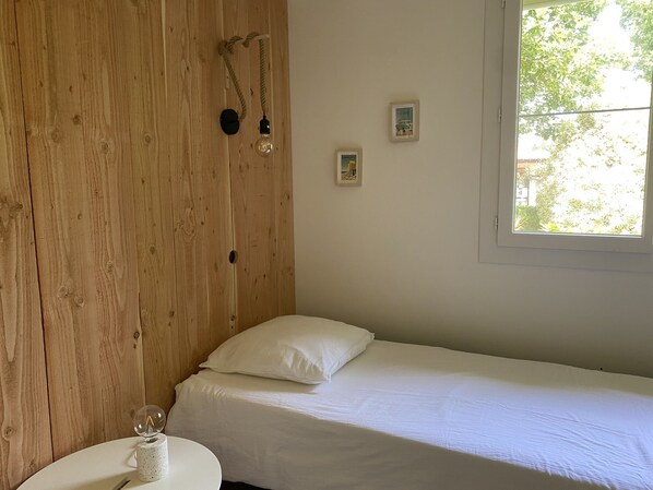 A room with a wooden wall, a bed with white bedding, a small round table with a lamp, and a window with a view of trees. - A one-time special offer for the month of September: a magnificent villa in the Landes style (Messanges)