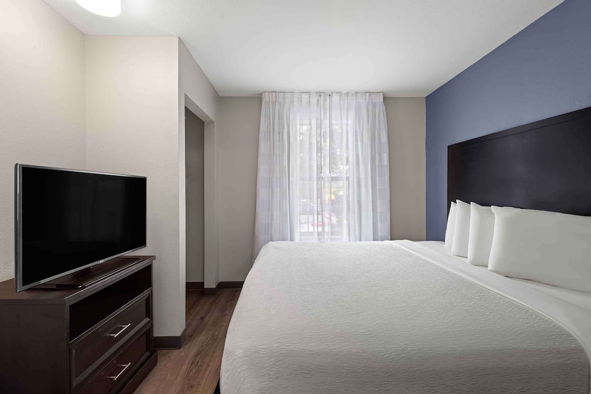 Una habitación de hotel moderna con una cama grande, un televisor de pantalla plana sobre un mueble de madera y vistas al exterior a través de cortinas traslúcidas.
