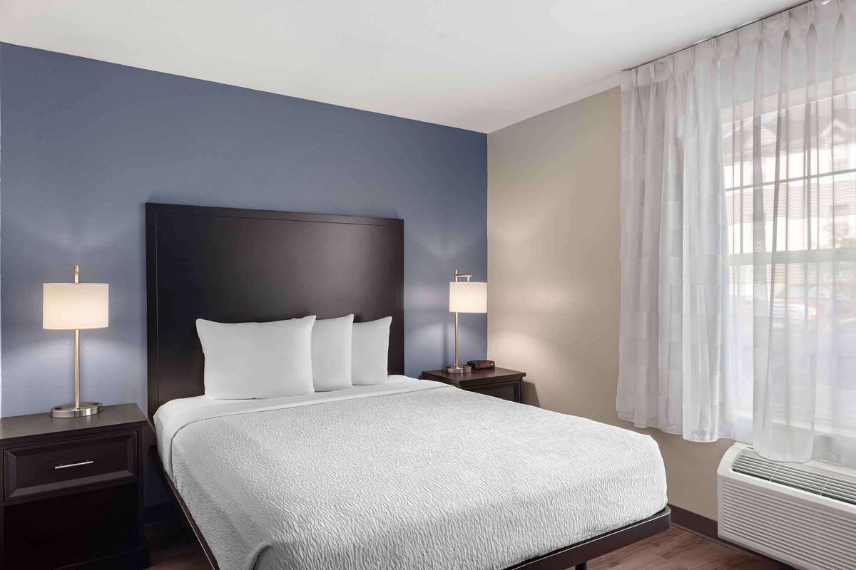 Habitación de hotel con una cama grande, dos mesitas de noche, una ventana con cortinas translúcidas y un equipo de aire acondicionado.