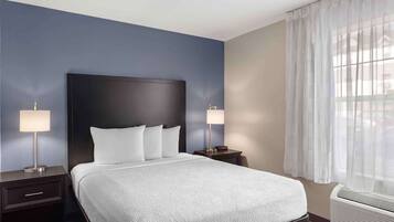 A hotel room with a large bed, two bedside tables, a nightstand, a window with sheer curtains, and an air conditioning unit.
