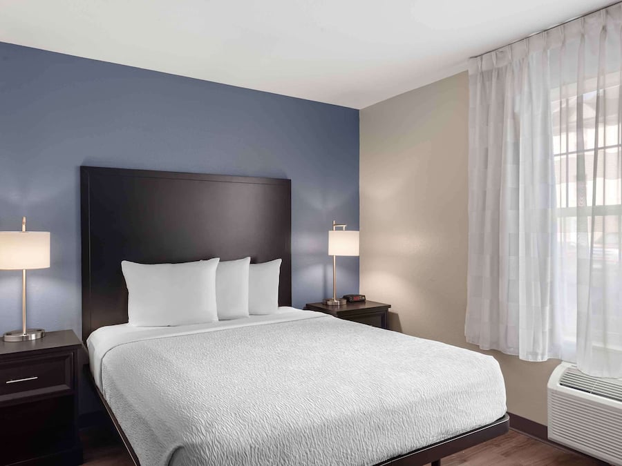 A hotel room with a large bed, two bedside tables, a nightstand, a window with sheer curtains, and an air conditioning unit.