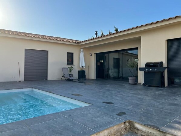 A swimming pool with a tiled deck, a barbecue grill, and a covered patio area.