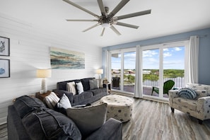 A spacious living room with a ceiling fan, a large window with a view of a body of water, and a balcony with outdoor furniture.