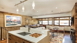 A modern kitchen with a large island, a dining area with a sofa and chairs, and a large window with a view of the mountains. - Fasching Haus Unit 6 | Condo with Beautiful Decor, Spacious Layout, Great Location (Aspen)