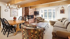 A cozy living room with a brick fireplace, wooden ceiling beams, a dining area with a wooden table and chairs, and a large window providing natural light.