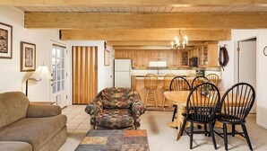 A cozy living room with a sofa, a patterned armchair, a dining table with chairs, a kitchen in the background, and a wooden ceiling.