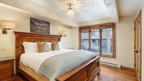 Une chambre à coucher avec un grand lit, une tête de lit en bois, deux lampes de chevet, un lustre, un tableau au mur et une fenêtre donnant sur l’extérieur.