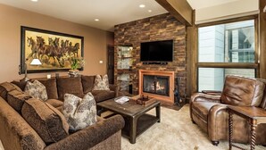 A living room with a fireplace, a large TV, and a painting of horses. - Durant Condominiums Unit C6 | Ski-In Condo, 2 Blocks from the Silver Queen Gondola (Aspen)
