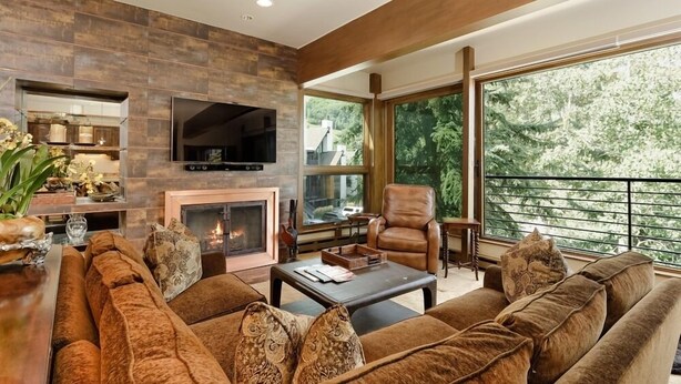 A modern living room with a fireplace, leather sofa, and a large window offering a view of trees. - Durant Condominiums Unit C6 | Ski-In Condo, 2 Blocks from the Silver Queen Gondola (Aspen)