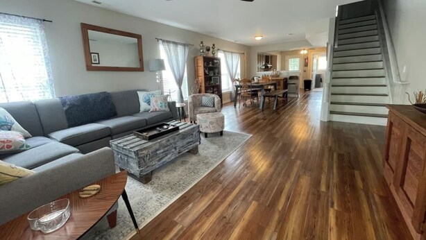 A spacious living room with a grey sofa, a coffee table, and a dining area with a table and chairs. A staircase leads to an upper level. - Chesapeake Chalet- Cozy,Close to Hard Rock and All (Atlantic City)