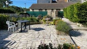 A stone patio with outdoor seating, a garden with various plants, and a building with a tiled roof in the background.