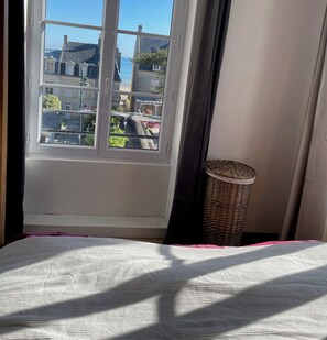 A room with a large window offering a view of buildings and a body of water. - Apartment La Vigie - 30 m from the sea, small sea view  (Saint-Aubin-sur-Mer)