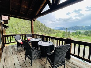 Une terrasse en bois, agrémentée de chaises en osier et d’une table ronde, offrant une vue sur un paysage montagneux avec un lac.