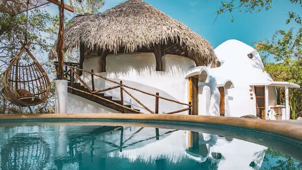 Ein Pool mit einer strohgedeckten Struktur und ein weißes Gebäude mit einem einzigartigen architektonischen Design.