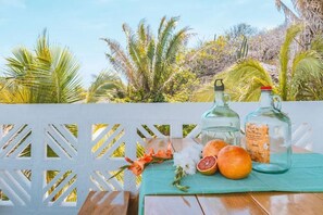 Ein Holztisch mit grüner Tischdecke, zwei Flaschen mit Etiketten, eine halbiertes Grapefruit und einige Orangen auf einem Balkon mit weißem Gittergeländer und tropischen Pflanzen im Hintergrund.