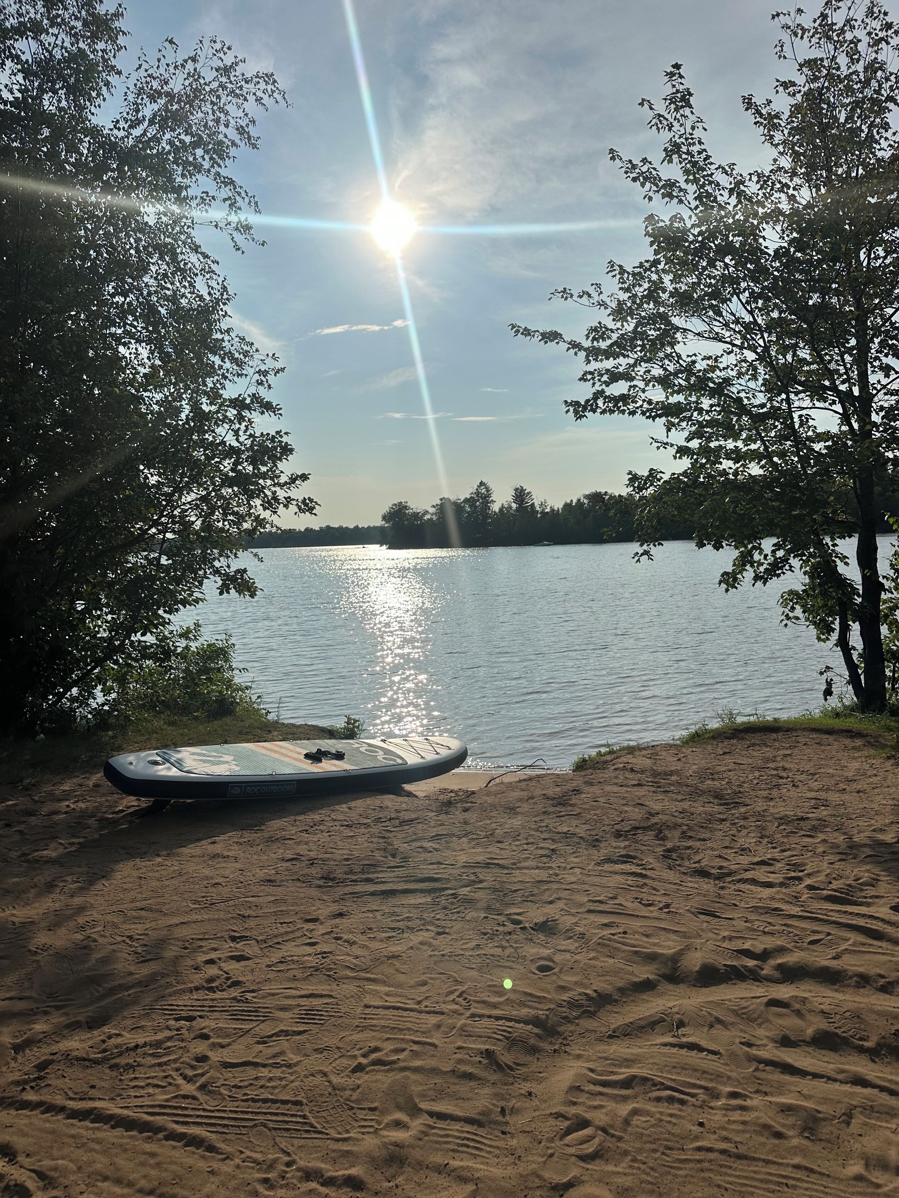 A sandy beach with a paddleboard and a calm lake in the background.