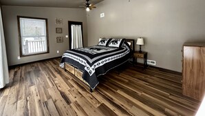 A bedroom with a large bed, a ceiling fan, a window with blinds, and a wooden floor.