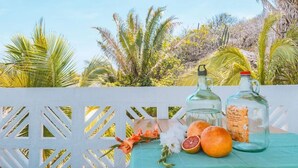 A wooden table with a green cloth, two glass bottles with labels, a half-cut grapefruit, and a few oranges on a balcony with a white lattice railing and tropical plants in the background. - Remote Beach Eco-Dome w/pool. Near Roca Blanca (Oax.)