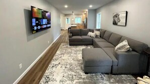 Un salon moderne avec un canapé d’angle, un téléviseur à écran plat fixé au mur et une cuisine en arrière-plan.
