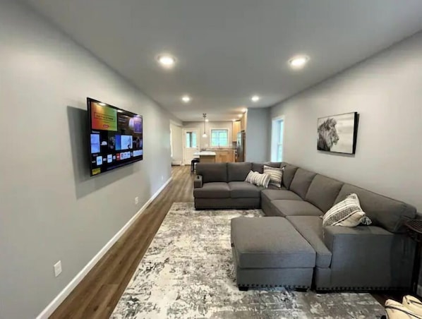 A modern living room with a sectional sofa, a flat-screen TV mounted on the wall, and a kitchen in the background. - Private New House | Food + Bars + Downtown (Winooski)