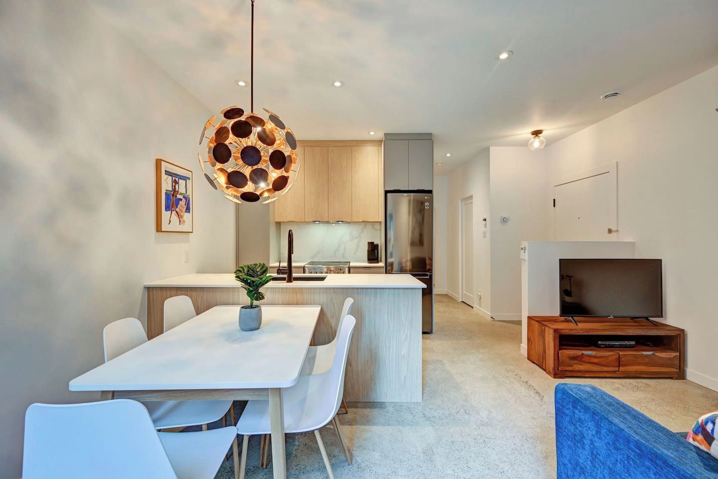 A modern kitchen with a central island, white dining chairs, a hanging light fixture, and a flat-screen TV in a wooden entertainment unit.